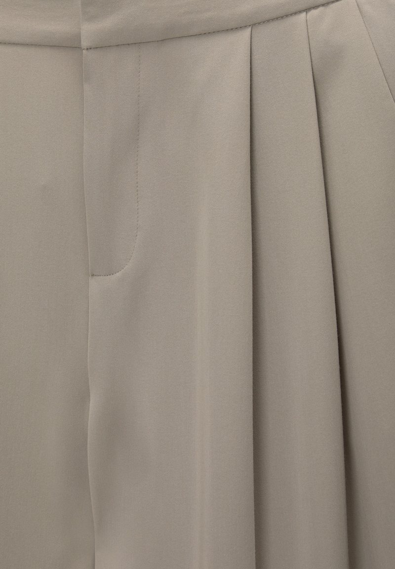 Tissu beige clair avec une texture douce, présentant des plis et une poche latérale. Design cintré adapté à une tenue formelle ou décontractée.