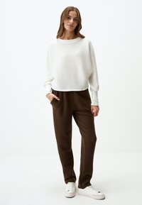 Weißer, strukturierter Strickpullover mit rundem Halsausschnitt und tiefen Schultern; kombiniert mit dunkelbraunen, glatten Stoff-Jogginghosen und weißen Turnschuhen.