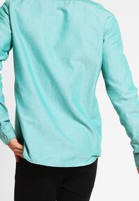 Hell-türkisfarbenes Langarmhemd aus einem glatten Stoff. Es hat eine lockere Passform, geknöpfte Manschetten und einen abgerundeten Saum hinten.