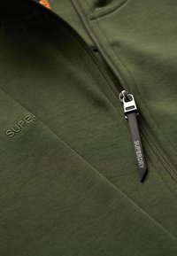 Grøn zip-up jakke lavet af et blødt stof. Har en sølv lynlås med et sort håndtag mærket "SUPERDRY" og broderet logo i lysere grøn.