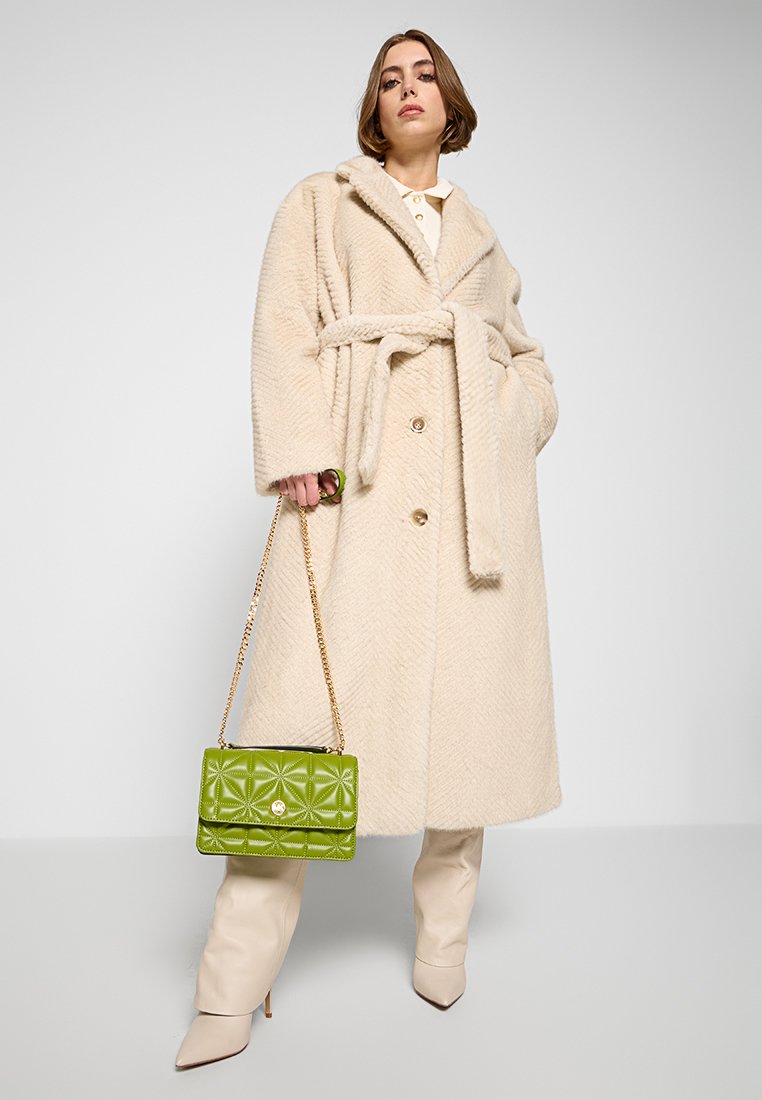 Beige vissengraatjas met een strikriem, voorzien van grote knopen; draagt een groene gewatteerde handtas met een gouden sluiting en kettingriem.