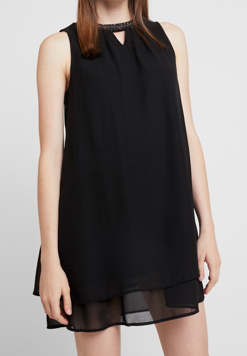 Robe noire sans manches avec détail de col perlée, découpe en forme de serrure et ourlet transparent superposé portée par une personne debout sur fond uni.