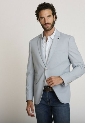Homme aux cheveux bouclés et à la barbe, portant un blazer gris clair sur une chemise blanche à motifs et un jean bleu, sur fond uni.