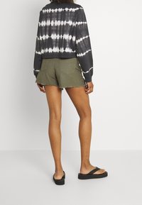 Top de manga larga en blanco y negro con estampado de tie-dye, combinado con pantalones cortos de mezclilla verde oliva con bordes deshilachados. Sandalias planas negras completan el look.