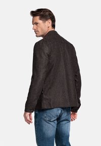 Donkergrijze wollen jas van een wolmix met een gestructureerde kraag, knoopsluiting en twee zijzakken. Gekleed over een blauwe spijkerbroek, met zicht op de achterkant.