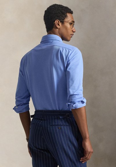 Άνδρας με γαλάζιο πουκάμισο με ρυχώσεις στα μανίκια και παντελόνι σκούρου μπλε με ρίγες, που κοιτάζει πλάγια μπροστά σε ουδέτερο φόντο.