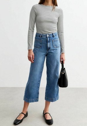 Frau trägt ein gestreiftes Langarmshirt, hochtaillierte, weite, gekürzte Jeans, schwarze Ballerinas und hält eine schwarze Handtasche.