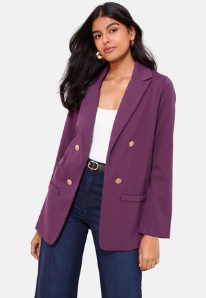 Vrouw draagt een paarse double-breasted blazer met gouden knopen, een wit top, donkerblauwe spijkerbroek en gouden creolen tegen een effen achtergrond.