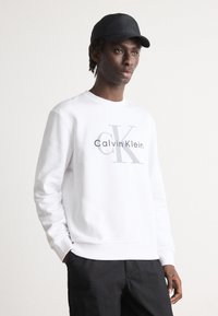 Felpa bianca con logo frontale che presenta "Calvin Klein" e "CK" in grigio. Collo rotondo, maniche lunghe e polsini a costine. Indossata con pantaloni neri.
