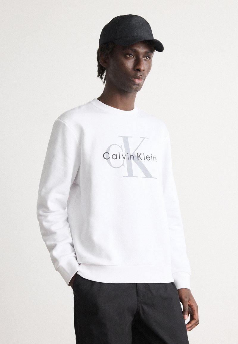 Felpa bianca con logo frontale che presenta "Calvin Klein" e "CK" in grigio. Collo rotondo, maniche lunghe e polsini a costine. Indossata con pantaloni neri.