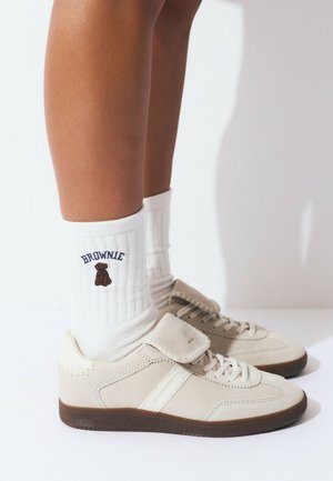 Zapatillas de ante beige con rayas blancas y una correa de Velcro, combinadas con calcetines blancos de canalé que presentan un oso marrón y bordado "BROWNIE".