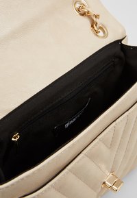 Beige Lederhandtasche mit gestepptem Design, ausgestattet mit einem goldfarbenen Kettendetail, schwarzem Stofffutter und einer Innenzippertasche.