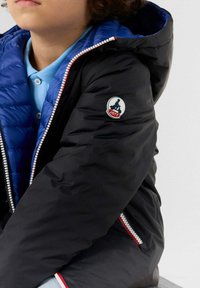 Giacca a vento nera con fodera interna blu e dettagli in cerniera rossa. Presenta una toppa con logo rotondo sul braccio sinistro. Texture liscia.