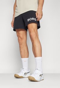 Juodi šortai su baltais užrašu "BORG", derinami su baltais sportiniais kojiniais ir baltais sportiniais bateliais su smėlio spalvos padu ir juodais akcentais.