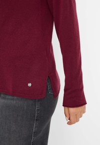 Maglione in maglia bordeaux con maniche lunghe, polsini a coste, fessure laterali e un accento metallico vicino all'orlo. Texture morbida, design casual.