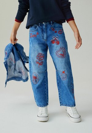 Child wearing blue jeans with red embroidered designs including roses, hearts, cherries, and cupcake, holding a matching denim jacket.