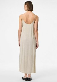 Robe maxi en lin beige avec un dos décolleté, des bretelles fines et une silhouette droite. Des chaussures noires plates complètent la tenue.