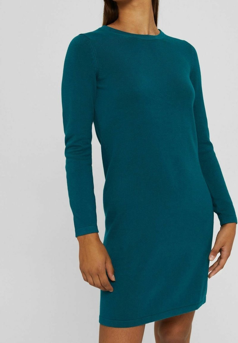 Femme portant une robe tricotée ajustée à manches longues de couleur sarcelle, debout devant un fond gris clair uni.