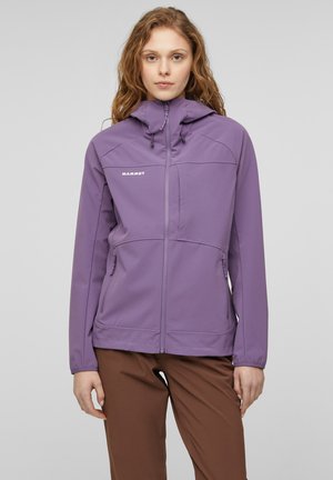 Giovane con capelli mossi che indossa una giacca con cappuccio viola con cerniera frontale e pantaloni marroni, in piedi su uno sfondo uniforme.