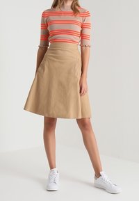 Falda en línea A de color beige con bolsillos laterales y una textura suave, combinada con una blusa de manga corta a rayas en beige y naranja neón, usada con zapatillas blancas.