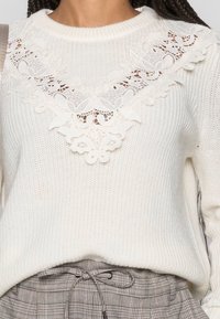 Pull en maille blanche avec un détail en dentelle complexe en forme de V au niveau de l'encolure. Texture côtelée avec des manches longues.