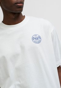 White short-sleeve T-shirt with a round neck and a blue embroidered circular logo featuring a bee and laurel leaves. Soft cotton material.