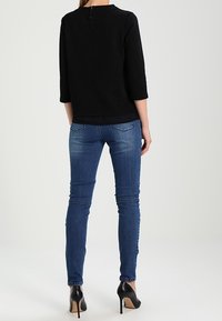 Sort tekstureret top med trekvartærmer og lynlås i ryggen, parret med blå slim-fit jeans og sorte højhælede sko.