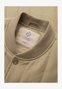 Chaqueta acolchada beige con cuello de canalé y botones de presión, mostrando etiqueta blanca con la marca "Ombre Heritage" y talla S/M en el interior del escote.