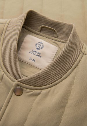 Beżowa pikowana kurtka z prążkowanym kołnierzem i zatrzaskiem, z widoczną białą metką z nazwą marki "Ombre Heritage" oraz rozmiarem S/M wewnątrz kołnierza.