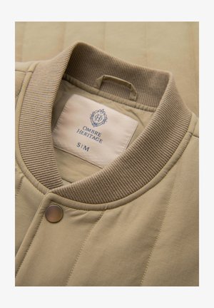 Beżowa pikowana kurtka z prążkowanym kołnierzem i zatrzaskiem, z widoczną białą metką z nazwą marki "Ombre Heritage" oraz rozmiarem S/M wewnątrz kołnierza.