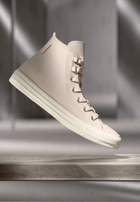 Baskets montantes en toile beige avec une semelle et des lacets en caoutchouc blancs, présentées en lévitation au-dessus d'une surface en pierre dans un éclairage doux.