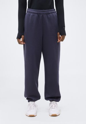 Άτομο που φοράει σκούρο μπλε φόρμα με ελαστική μέση και μανσέτες, συνδυασμένη με λευκά αθλητικά παπούτσια και μαύρο μακρυμάνικο τοπ.