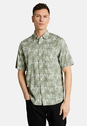 Mann trägt kurzärmliges Hemd mit grünem Palmenmuster, steht mit einer Hand in der Hosentasche vor einfarbigem Hintergrund.