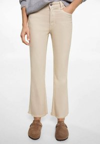 Beige flared jeans with a raw hem, featuring a front pocket and metal button closure; worn with brown suede clogs.