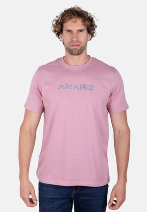 Hombre con el pelo rizado, vestido con una camiseta rosa claro con el texto "AMARO" y vaqueros azules, de pie frente a un fondo liso y claro.