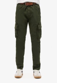 Pantalones cargo verde oliva con bolsillos laterales, cinturón de cuero marrón y zapatillas blancas con detalles negros sobre fondo blanco.