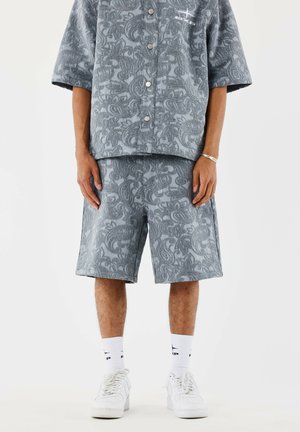 Hombre vistiendo una camisa de manga corta y pantalones cortos a juego con estampado de paisley gris, con calcetines blancos y zapatillas deportivas contra un fondo blanco.