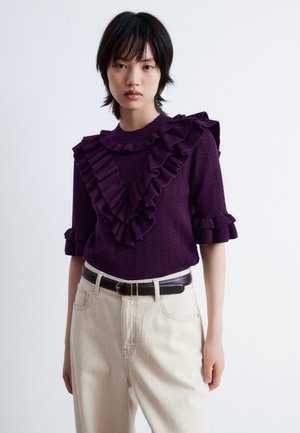 Purple knitted sweater featuring ruffled accents along the neckline and sleeves, paired with beige high-waisted pants and a brown belt.