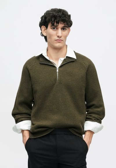 Olivgrüner Strickpullover mit einem Viertelreißverschluss und geripptem Design, über einem weißen Hemd mit Kragen getragen. Schwarze Hose.