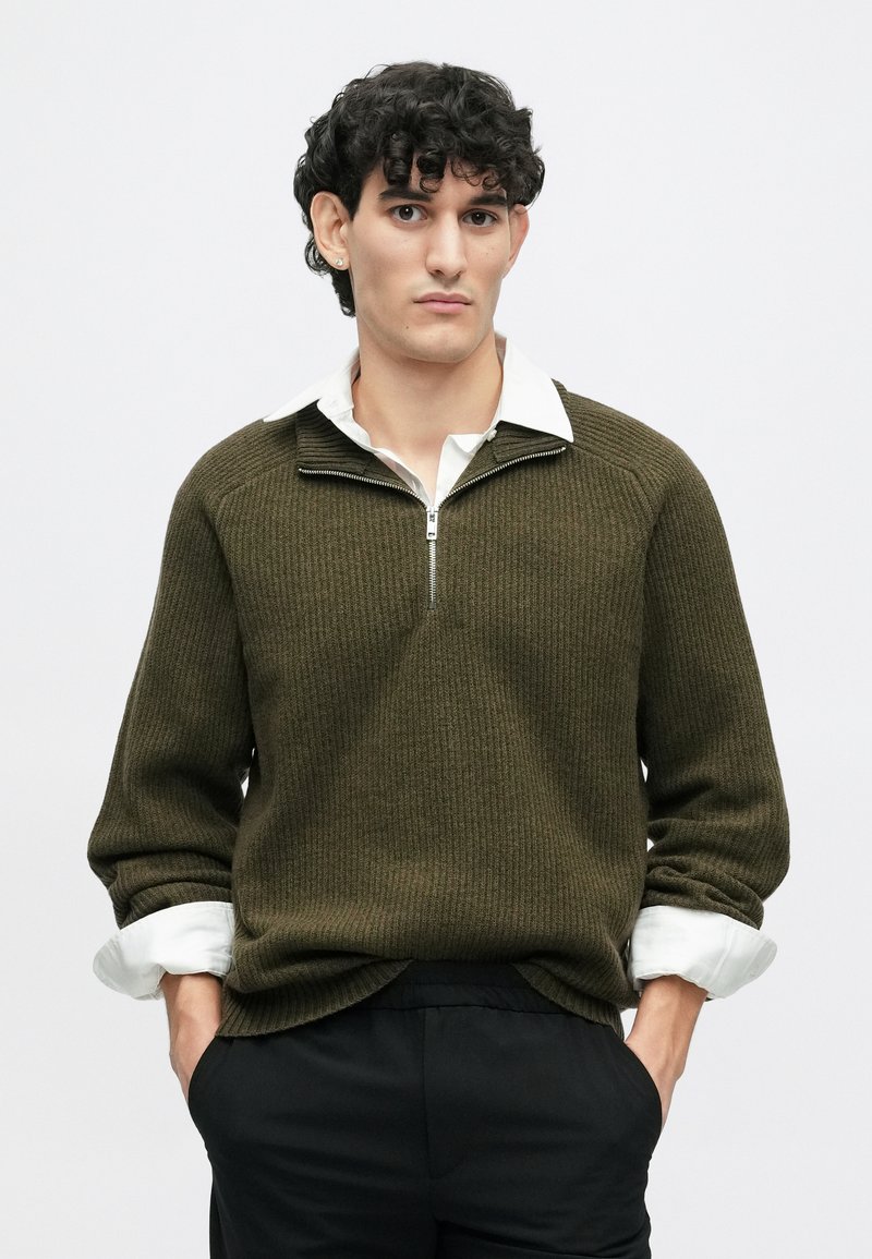 Olivengrøn strikket sweater med en kvart lynlås og ribbet tekstur, lagket over en hvid skjorte med krave. Sorte bukser.