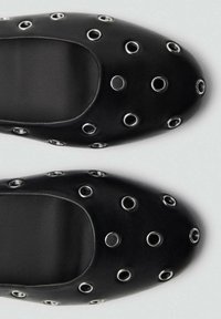 Schwarze Schuhe mit einer glatten Lederoberfläche, ausgestattet mit großen metallischen Ösen, die gleichmäßig über die Oberseite verteilt sind. Abgerundete Zehenform.