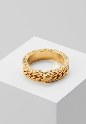 Bague en or avec un motif clé grecque et un design en chaîne avec un emblème de tête de lion, posée sur une surface blanche.