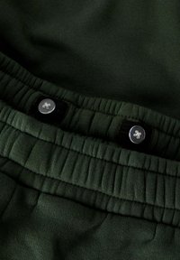 Mørkegrønne sweats med et blødt, struktureret stof, plisseret talje, elastiske detaljer og to sorte knapper foroven.