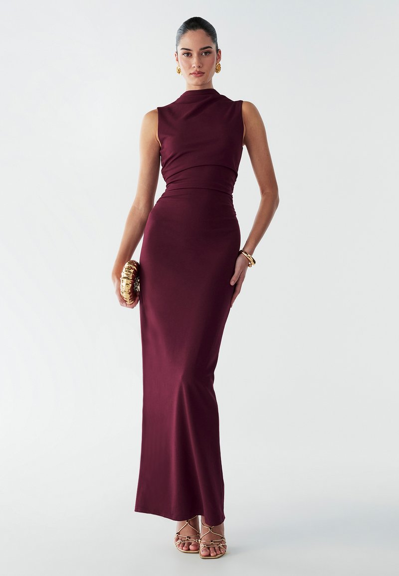 Burgundijas bez piedurknēm vakartērps ar pieguļošu siluetu, augstu kakla izgriezumu un savilktām auduma detaļām. Kombinēts ar aksesuāriem ar zelta akcentiem un sandālēm.