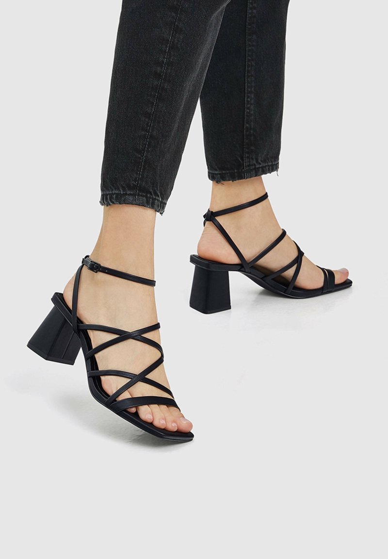 Pieds portant des sandales noires à talons blocs avec lanières, associées à un jean noir raccourci sur un fond gris clair.