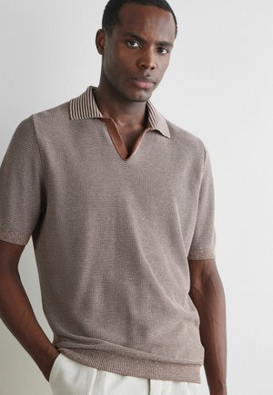 Mann trägt ein kurzärmeliges, gestricktes Polo mit dezentem braunem und beigem Muster und steht vor einer einfarbigen hellgrauen Wand.