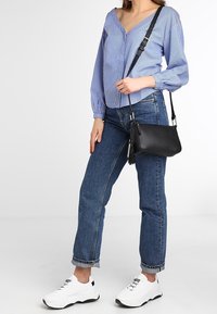 Woman wearing blue striped blouse, blue rolled-up jeans, white sneakers, and carrying a black crossbody bag with tassel detail.