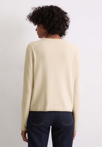 Heller cremefarbener Pullover mit langen Ärmeln, Rundhalsausschnitt und geripptem Saum; kombiniert mit dunkelblauen Jeans. Weicher Stoff, minimales Design.