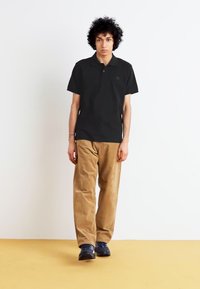 Black short-sleeve polo shirt paired with light brown corduroy pants and dark sneakers, standing against a white background on a yellow floor.