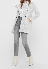 Abrigo gris con textura, cuello alto, doble botonadura y bolsillos laterales, combinado con jeans grises y botines blancos con tacón.